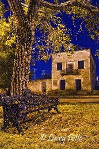 Cottonwood tree and park bench on the Roma plaza in early evening. A sandstone historic building on the west side of the plaza awaits restoration.